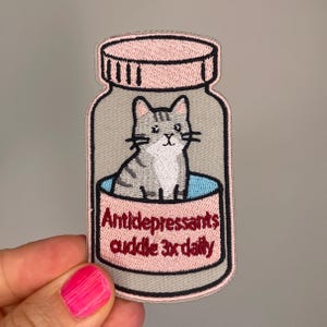 Könnte beinhalten: Gestickter Aufnäher mit einer grauen getigerten Katze in einer rosa Pillenflasche. Die Flasche hat einen rosa Deckel und den Text "Antidepressiva kuscheln 3x täglich" in Burgunderrot.