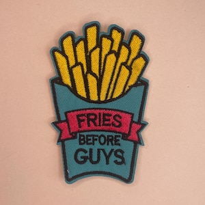 May include: Embroidered patch with a teal container of golden french fries. A red banner across the container reads "FRIES BEFORE GUYS" in white text. The patch has a black border, and is a fun accessory.