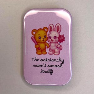 Könnte beinhalten: Rechteckige, hellviolette Anstecknadel mit abgerundeten Ecken. Sie zeigt einen Cartoon-Bären mit einem Baseballschläger und ein Kaninchen mit einem Stachelball. Der Text "The patriarchy won't smash itself!" steht darunter.