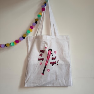 Könnte beinhalten: Eine weiße Tragetasche mit einer Grafik eines rosa Baseballschlägers mit Gesicht, schwarzen Haaren und einer Schleife. Der Text Full of Love and rage! ist von kleinen rosa Herzen umgeben. Die Tasche hat zwei weiße Tragegriffe und eine rechteckige Form.
