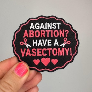 May include: A black embroidered patch with a scalloped edge and pink border. The text reads AGAINST ABORTION? HAVE A VASECTOMY! in white and pink lettering. Two small pink scissors icons are placed between the lines, and three pink hearts are below.