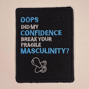 Pode incluir: Patch bordado preto com o texto "OOPS DID MY CONFIDENCE BREAK YOUR FRAGILE MASCULINITY?" em branco e azul. Um gráfico de chupeta branca na parte inferior. O patch é retangular com cantos arredondados.