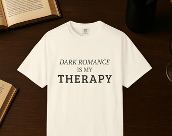 May include: Cream-colored t-shirt with the text "DARK ROMANCE IS MY THERAPY" in black. The shirt is displayed on a dark wooden surface, alongside open books, a wine bottle, and a wine glass. The phrase "COLOR MAY VARY SLIGHTLY DUE TO LIGHTING OR SCREEN SETTINGS" is at the bottom.