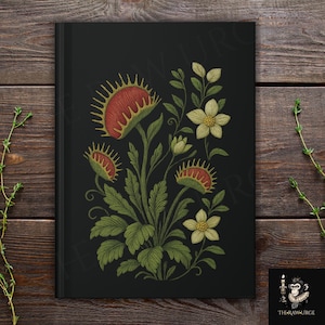 Op de afbeelding: Zwart notitieboek met een geborduurd ontwerp van Venus vliegenvallen en bloemen in rood, groen en crème. De cover art is een gedetailleerde botanische illustratie. Het notitieboek staat tegen een donkere houtnerf achtergrond.