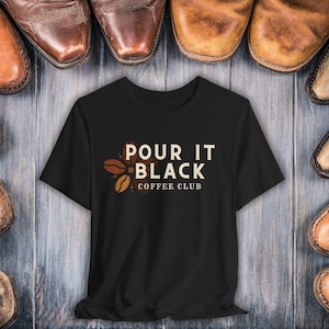 May include: Black t-shirt with the text "POUR IT BLACK COFFEE CLUB" in white and brown. The design includes coffee beans. The shirt is displayed on a wooden surface, surrounded by brown leather shoes.