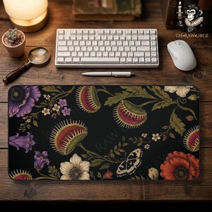 May include: A rectangular desk mat featuring a black background with a botanical pattern of purple and red flowers, green Venus flytraps, and a butterfly. The design includes intricate foliage and small white blossoms across the surface.