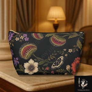 May include: A black zippered pouch featuring a botanical print of green and red Venus flytraps, white and purple flowers, and a butterfly. The design includes the text THE RAW URGE in a subtle repeating pattern across the dark fabric surface.