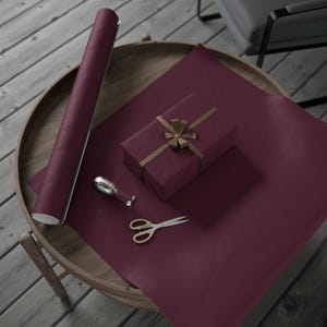 May include: A rectangular gift box wrapped in deep burgundy paper with a gold ribbon bow, accompanied by a matching roll of wrapping paper, a small silver tape dispenser, and a pair of metal scissors with gold-toned handles.