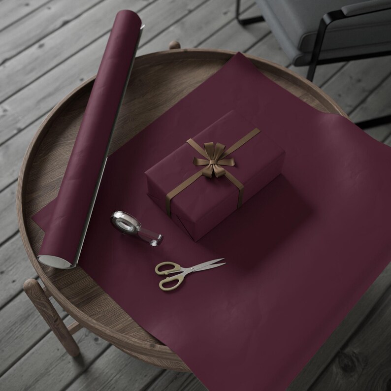 May include: A rectangular gift box wrapped in deep burgundy paper with a gold ribbon bow, accompanied by a matching roll of wrapping paper, a small silver tape dispenser, and a pair of metal scissors with gold-toned handles.