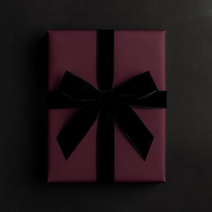 May include: A rectangular gift box wrapped in deep burgundy paper, featuring a wide, black velvet ribbon tied in a large, centered bow. The ribbon runs vertically and horizontally across the surface, creating a structured and elegant presentation.
