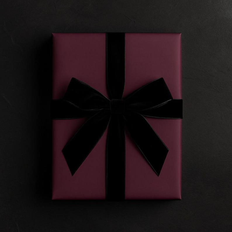 May include: A rectangular gift box wrapped in deep burgundy paper, featuring a wide, black velvet ribbon tied in a large, centered bow. The ribbon runs vertically and horizontally across the surface, creating a structured and elegant presentation.