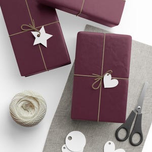 May include: Three rectangular gifts wrapped in deep burgundy paper, tied with thin tan twine. One gift features a white star-shaped tag, another a white heart-shaped tag. A ball of cream-colored twine and black scissors are also present.