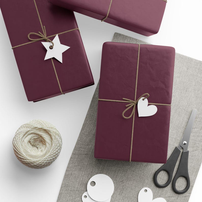 May include: Three rectangular gifts wrapped in deep burgundy paper, tied with thin tan twine. One gift features a white star-shaped tag, another a white heart-shaped tag. A ball of cream-colored twine and black scissors are also present.