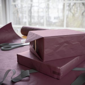 May include: Two rectangular boxes wrapped in deep plum-colored paper with a textured, slightly wrinkled finish. A roll of matching wrapping paper lies nearby, along with a pair of grey scissors and a length of grey ribbon on the surface.
