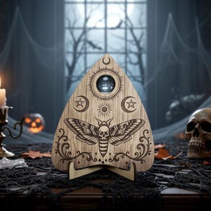 May include: A wooden Ouija board with a moth and skull design, featuring a planchette. The board has celestial symbols and is set against a spooky Halloween backdrop with a skull and cobwebs.