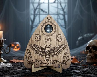 Könnte beinhalten: Ein hölzernes Ouija-Brett mit einem Motten- und Totenkopf-Design, mit einer Planchette. Das Brett hat himmlische Symbole und ist vor einem gruseligen Halloween-Hintergrund mit einem Totenkopf und Spinnweben platziert.