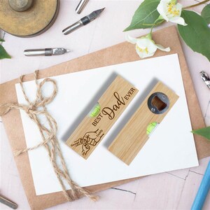 Op de afbeelding: Twee houten waterpassen, &eacute;&eacute;n met een flesopener, beide gegraveerd met "Best Dad Ever". Elk heeft een kleine waterpas en gepersonaliseerde namen. De gereedschappen zijn lichtbruin en liggen op een wit oppervlak.