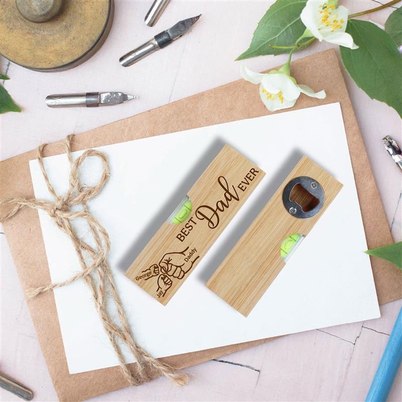 Op de afbeelding: Twee houten waterpassen, &eacute;&eacute;n met een flesopener, beide gegraveerd met "Best Dad Ever". Elk heeft een kleine waterpas en gepersonaliseerde namen. De gereedschappen zijn lichtbruin en liggen op een wit oppervlak.