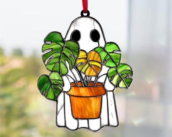May include: A stained glass ghost-shaped ornament holding a potted plant. The ghost is white with black eyes, and the plant has green and yellow leaves in an orange pot. A red ribbon is attached for hanging.