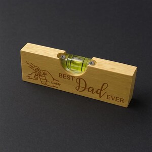 Op de afbeelding: Een houten waterpas met de tekst "BEST Dad EVER" gegraveerd. Het heeft een heldere buis met groene vloeistof en twee schroeven. De waterpas heeft ook een illustratie van twee handen en de namen "George" en "Jay".