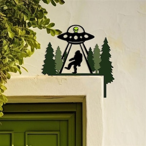 May include: A dark green metal wall art piece depicting a UFO with an alien inside, a silhouette of Bigfoot, and pine trees. The UFO has a green alien head in the center. The art piece is mounted on a white wall.