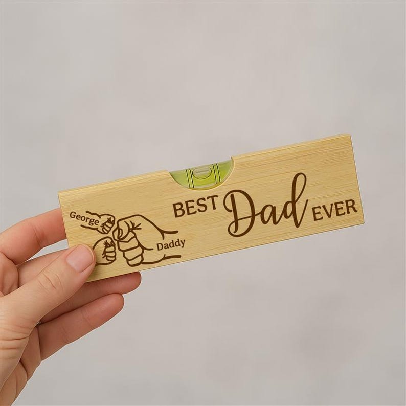Op de afbeelding: Een houten waterpas met de tekst "BEST Dad EVER" en een afbeelding van twee handen die elkaar vasthouden. De naam "George" staat boven &eacute;&eacute;n hand en "Daddy" onder de andere. Het heeft een heldere waterpas.