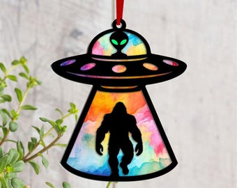 May include: Black UFO-shaped ornament with a colorful watercolor background featuring an alien and a silhouette of Bigfoot. The ornament is suspended by a red ribbon, perfect for holiday decor or a unique gift.