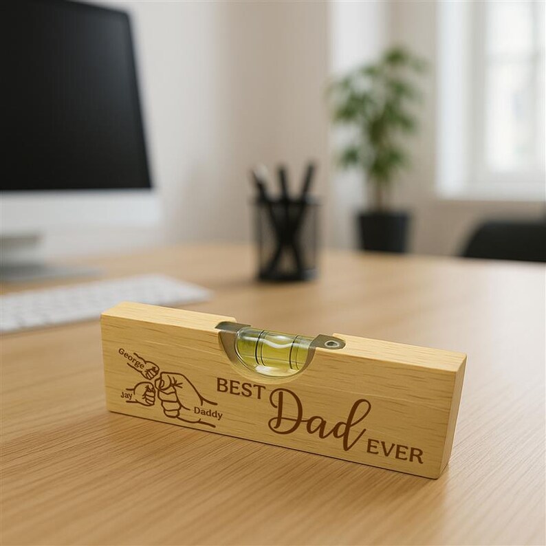 Op de afbeelding: Een lichtbruin houten waterpas met een luchtbel, met de tekst "BEST Dad EVER" en een afbeelding van handen die elkaar vasthouden. Het waterpas ligt op een houten oppervlak.