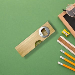 Op de afbeelding: Een houten flesopener met een metalen flesopener en een waterpas. De opener wordt omringd door kantoorbenodigdheden, waaronder een klein schoolbord, potloden en een liniaal, op een groene achtergrond.