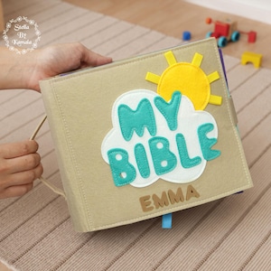May include: A beige felt book with a carrying handle, featuring a white cloud applique with the teal text "MY BIBLE" and a yellow sun above it. The name "EMMA" is stitched in brown felt letters at the bottom of the cover.