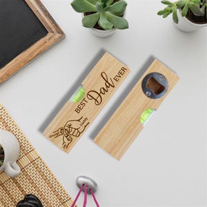 Op de afbeelding: Twee houten flesopeners met een waterpas en de tekst "Best Dad Ever" gegraveerd. E&eacute;n opener heeft een flesopener aan het uiteinde, de andere heeft een waterpas. De openers zijn lichtbruin en hebben een groene waterpasbel.