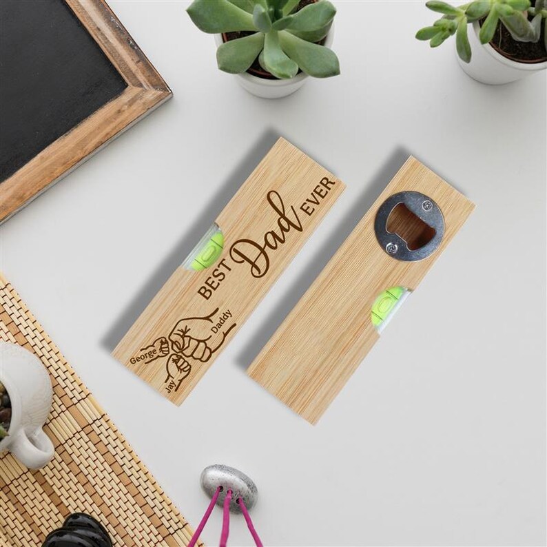 Op de afbeelding: Twee houten flesopeners met een waterpas en de tekst "Best Dad Ever" gegraveerd. E&eacute;n opener heeft een flesopener aan het uiteinde, de andere heeft een waterpas. De openers zijn lichtbruin en hebben een groene waterpasbel.