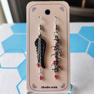 Puede incluir: Un par de pendientes asimétricos en una tarjeta beige. Uno presenta un ala negra con cadenas y cuentas rojas. El otro tiene un diseño en espiral plateado con cuentas rojas. La tarjeta tiene el texto "studio.wyy".