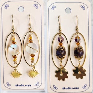Puede incluir: Dos pares de pendientes hechos a mano. Un par presenta un marco ovalado dorado con un orbe blanco, alambre dorado y un dije de sol. El otro par tiene un marco ovalado dorado con una cuenta morada, alambre dorado y un dije de engranaje. El texto "studio.wyy" es visible.