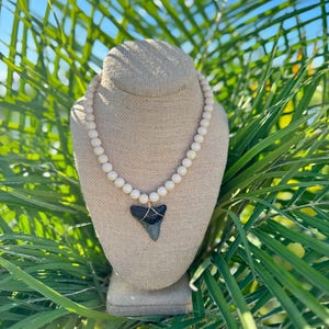 May include: A necklace featuring a shark tooth pendant, wire-wrapped in gold, suspended from a strand of light-colored wooden beads. The necklace is displayed on a beige jewelry bust against a backdrop of green foliage and blue sky.