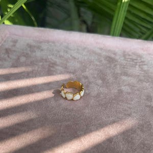 以下が含まれることがあります： 白いハート型のアクセントが付いたゴールドカラーのリング。リングはピンク色の背景に置かれ、日光によって影ができています。このリングはジュエリーです。