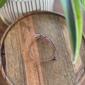 May include: A colorful woven bracelet with a small black charm. The bracelet features a braided design with pink, yellow, blue, and green threads. It is displayed on a round wooden tray, with a plant in the background.