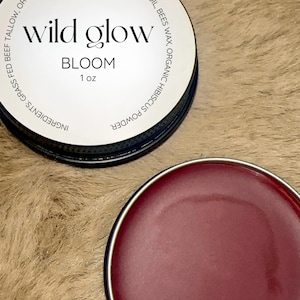 May include: A small, round tin container with a white label that reads "wild glow BLOOM 1 oz". The label lists ingredients including grass-fed beef tallow, organic castor oil, organic jojoba oil, bees wax, organic hibiscus powder. The tin is open and the inside is filled with a deep red balm.