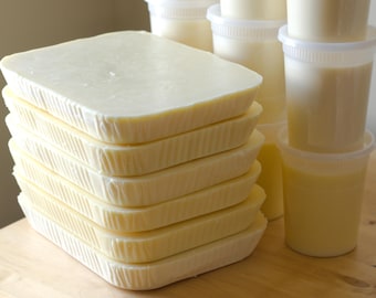 May include: A stack of rectangular, light yellow soap bars with textured edges. Several clear plastic containers filled with a creamy, pale yellow liquid are also visible, suggesting a handmade soap or cosmetic product.
