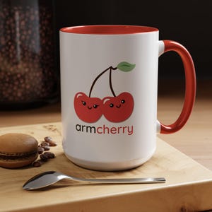 May include: White ceramic mug with a red rim and handle. The mug features a cartoon image of two cherries with faces, and the text "armcherry".