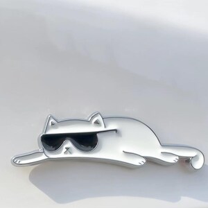 May include: A silver cat emblem wearing black sunglasses, in a relaxed pose. The emblem has a shiny surface. The text "InPalette" is in the upper left corner. The cat's design is modern and stylish.