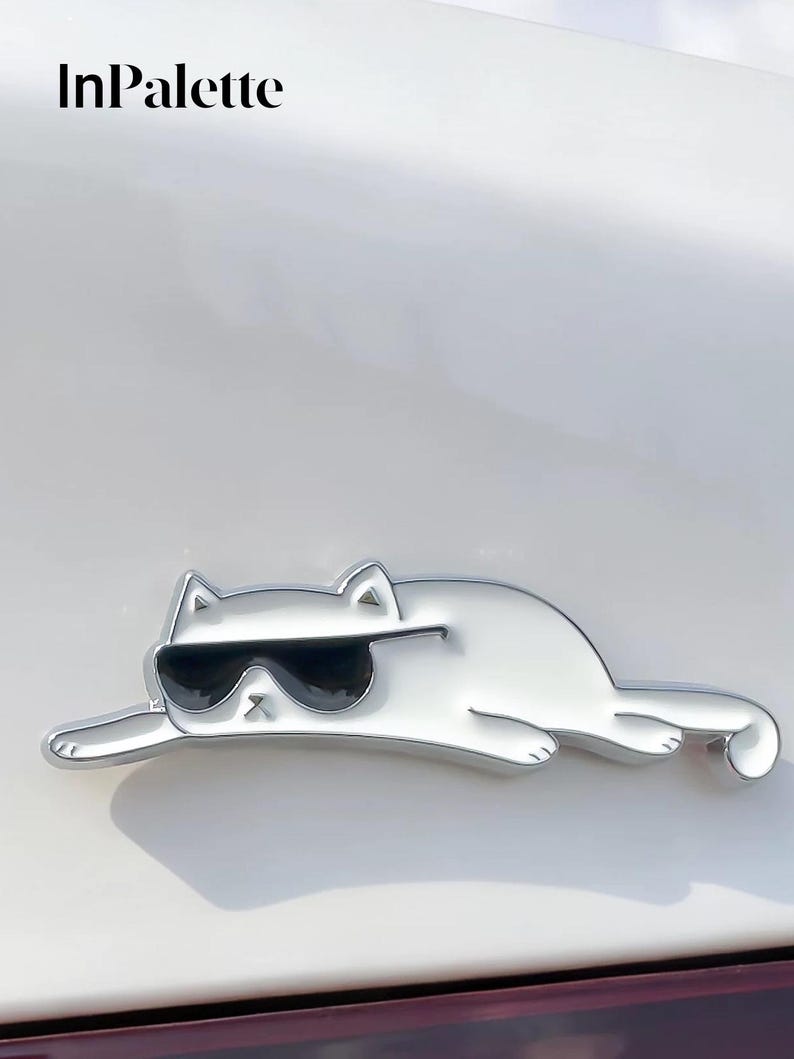 May include: A silver cat emblem wearing black sunglasses, in a relaxed pose. The emblem has a shiny surface. The text "InPalette" is in the upper left corner. The cat's design is modern and stylish.