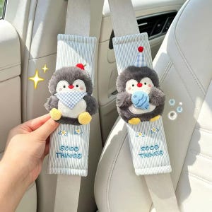 May include: Two light blue and white striped seat belt covers featuring plush grey penguin characters. One penguin wears a red hat and blue checkered scarf, the other a blue checkered party hat holding a blue fish. Both have embroidered flowers and the text GOOD THINGS.