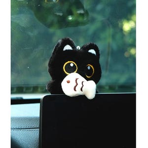 May include: A black plush cat with large yellow eyes and a white fish in its mouth. The cat has white accents on its ears and is hanging from a small loop. The cat is a car accessory.