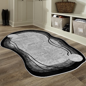 Puede incluir: Una alfombra abstracta con una forma única y una combinación de colores en escala de grises. La alfombra presenta un diseño en capas con tonos de gris y negro, delineado con un borde negro y un borde blanco. La alfombra está colocada sobre un suelo de madera.