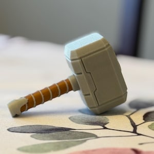 May include: A small, grey, plastic toy hammer with a brown handle. The hammer is shaped like Thor's hammer, Mjolnir.