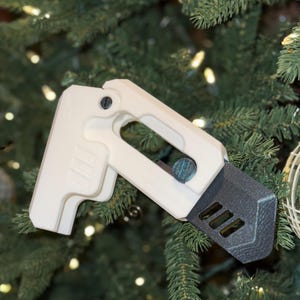 May include: A white plastic 3D printed ornament shaped like a gun, with a black and gray textured detail on the bottom. The ornament is hanging on a green Christmas tree.