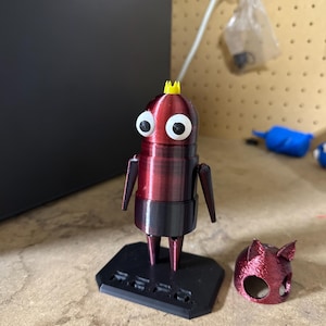 May include: A 3D printed red robot figurine with a yellow crown, large white eyes, and black arms and legs. The robot is standing on a black base with the word "REED" printed on it. A separate red 3D printed head piece is also visible.