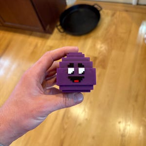 Puede incluir: Un personaje púrpura pixelado con una cara sonriente, sostenido en una mano. El personaje tiene ojos blancos y una boca roja. El diseño es en bloques y geométrico, que recuerda a los gráficos de videojuegos retro.