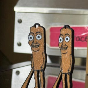 Puede incluir: Dos colgantes de personajes de dibujos animados marrones y brillantes con contornos negros. Cada colgante tiene una cara sonriente con ojos grandes y piernas largas. Los colgantes cuelgan de un pequeño agujero en la parte superior.